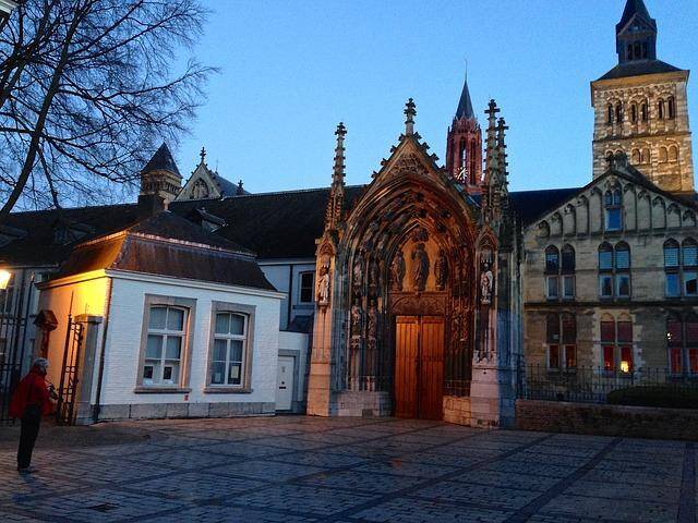 Schemeringszicht op de ingang van een gotische kerk met ingewikkelde architectuur, klaar voor een weekendje Limburg, omringd door andere historische gebouwen en een persoon die voorbij loopt.